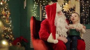 Noel ağacının yanında oturan genç bir çocuk Noel Baba ile konuşuyor. Parlak hediyeler ve şenlik ışıklarıyla çevrili..