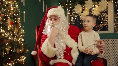 Noel Baba 'nın kucağında bir dilek tutan küçük çocuk, neşeli Noel ağacı arka planı, rahat koltuk, umut verici ifade, neşeli tatil