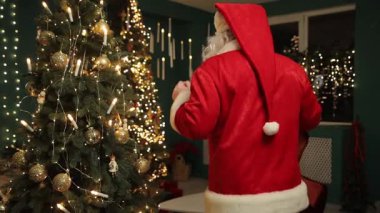 Noel Baba, sıcak tatil ışıklarıyla çevrili güzel dekore edilmiş bir ağaca altın süsler asar..