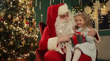 Noel Baba 'ya hediye veren gülümseyen kız, Noel ağacındaki parlak ışıklar, neşeli yüz ifadesi, şenlikli sandalye ayarları, samimi