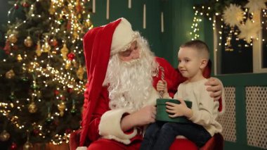 Neşeli genç bir çocuk, Noel Baba 'nın kucağında, sıcak Noel ışıkları ve hoş dekorasyonlarla otururken, bir tatil ziyafetinin tadını çıkarıyor..