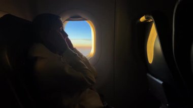 Yorgun kadın gezgin sabah uçuşu sırasında baş ağrısı nedeniyle uçak penceresinin yanında oturur. Yumuşak gün doğumu ışığı kabini doldurur. Yolculuk yorgunluğu ve duygusal yorgunluk gösterir..