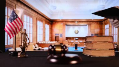 Ahşap tokmak, hukuk kitapları parlak mahkeme salonunda masanın üstünde. Arka planda görünen cübbeli yargıç otoriteyi, adaleti ve hukuki gücü simgeler..