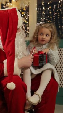 Noel hediyesini elinde tutan küçük kız Noel Baba 'nın yanında oturmuş bayram süslemeleri yapıyor. İçten bir aile anı..