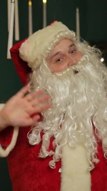 Noel Baba Noel ağacının önünde elini sallıyor, bayramlarda çocukları selamlıyor. Dikey video.