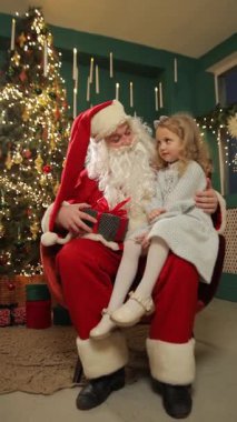 Noel Baba, süslü bir Noel ağacının yanında küçük bir kızı kucağında tutarak neşeli bir tatil anını paylaşıyor..