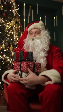 Noel Baba Noel hediyelerini süslenmiş bir Noel ağacının yanında otururken tutuyor..