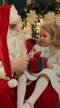 Küçük bir kız Noel Baba 'nın kucağında oturuyor, gülümsüyor ve Noel Baba ona parıldayan bir Noel ağacının önünde hediye paketini uzatıyor..