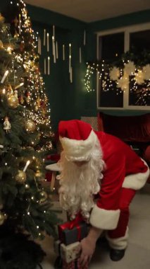 Noel Baba, Noel ağacının yanında diz çöker. Sıcak tatil ışıklarıyla dolu samimi bir festival odasında hediyeleri dikkatlice düzenler..