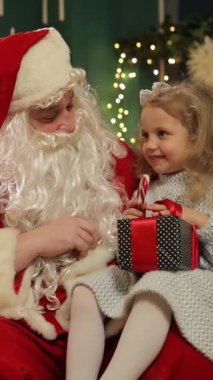 Noel Baba, elinde paketlenmiş bir hediye olan küçük bir kızın yanında oturur ve süslü bir Noel ağacının yanında samimi bir şekilde konuşur ve gülümserler..