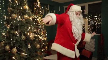 Noel Baba neşeli bir şekilde hareket eder ve ışıl ışıl bir Noel ağacının yanında dans eder. Arka planda sıcak, pırıl pırıl ışıklar ve neşeli bir tatil havası yaratır..