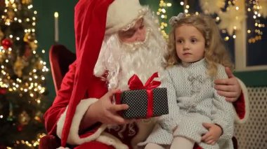 Noel Baba ve küçük bir kız Noel ışıklarının yanında otururlar ve sıcak bir bayram anında birbirlerine sarılı bir hediye tutarlar..