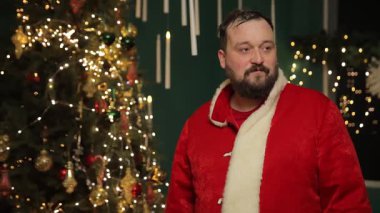 Bir adam Noel Baba kıyafetini ışık ve süslemelerle dolu bir festival odasında düzenleyerek Noel çekimine hazırlanır..