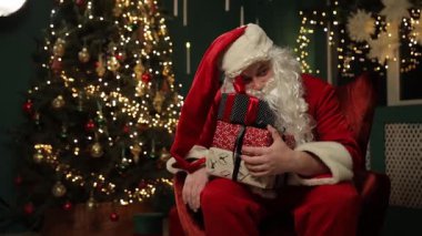 Noel Baba güzel süslenmiş bir Noel ağacının yanında otururken bir yığın hediyeyi neşeyle tutuyor..