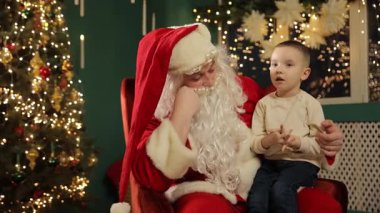 Noel Baba kucağında küçük bir çocukla oturur ve neşeli ışıklarla dolu sıcak bir Noel anını paylaşır..