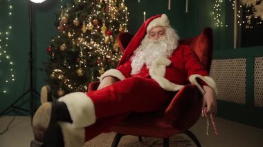Noel Baba, sıcak Noel ışıklarıyla çevrili bir bayram gecesinde gündelik kırmızı bir sandalyede oturur..