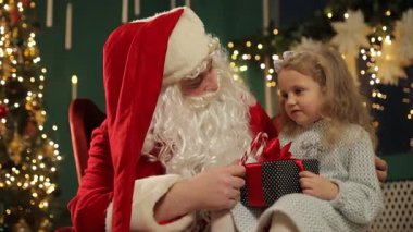 Noel Baba küçük bir kıza sarılı bir Noel hediyesi veriyor. Birlikte, ışıklarla süslenmiş sıcak bir tatil ortamında oturuyorlar..