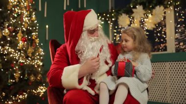 Noel Baba, gülümseyen küçük bir kıza şeker kamışı uzatıyor. Noel ağacının yanında, samimi bir festival odasında birlikte oturuyorlar.