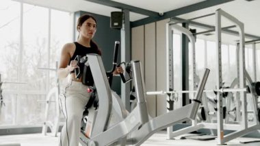 Kadın, geniş pencereli, ışık dolu bir spor salonunda eliptik bir eğitmen üzerinde egzersiz yapıyor. Koordineli kol ve bacak hareketi düşük etki yaratıyor..