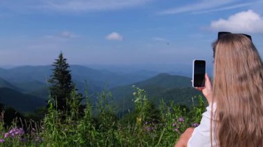 Uçsuz bucaksız bir dağ manzarasının önünde duran bir kadın, elinde telefonuyla katmanlı yeşil tepeleri ve mavi gökyüzünü fotoğraflıyor..