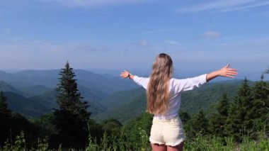 Bir kadın kollarını genişçe açar ve etrafındaki dağ manzarasının güzelliğinden zevk alır, özgür ve keyifli bir açık hava anı yaratır..