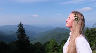 Bir kadın profilde gözleri kapalı duruyor, taze dağ havasını soluyor yeşil tepelere ve parlak mavi gökyüzüne karşı..