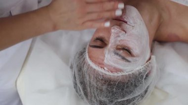 Bir müşteri yüzüne sürdüğü kalın beyaz bir maskeyle dinleniyor. Sakin spa ortamları ve yumuşak dokular rahatlatıcı bir cilt bakımı atmosferi yaratır..