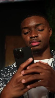 Afro-Amerikalı bir adam akıllı telefonunu kapalı mekanda hafif aydınlatılmış bir şekilde kontrol ediyor. Sıcak ışık yüzüne vurgu yapıyor, sakin ve odaklanmış bir yaşam tarzı yaratıyor..