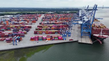 Rıhtım boyunca renkli nakliye konteynırları, vinçler ve kenetlenmiş gemilerle dolu yoğun bir kargo limanının yüksek açılı görüntüsü. Görüntüler aktif bir lojistik merkezi gösteriyor. 