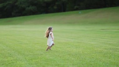 Küçük bir kız, yumuşak doğal ışıkla çevrili, geniş, açık otlak bir alanda yavaşça yürür. Huzurlu bir ortam, doğadaki sakin ve nazik bir çocukluk anını vurgular..