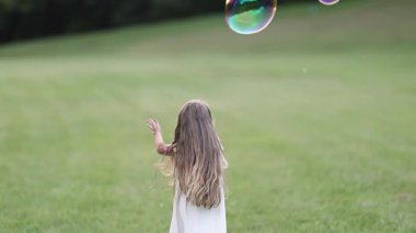Tatlı genç bir kız yeşil bir alanda duruyor yavaşça yüzen baloncuklara uzanıyor. Sessiz açık hava atmosferi ve yumuşak ışık sakin ve masum bir çocukluk anını vurgular..