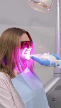 UV lambası ile diş beyazlatan ve koruyucu gözlük takan genç bir kadının dikey videosu. Modern bir diş kliniğinde diş beyazlatma
