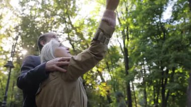 Aşık emekliler, ağaçların arasında gün ışığının arka planında birbirlerine sarılıyorlar. Yürüyüşteki mutlu kıdemli insanlar