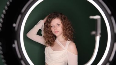 Kıvırcık saçlı genç bir kadın halka ışığının altında poz veriyor, saçlarına özgüvenle dokunuyor. Stüdyo estetiği, hafif ışıklandırma ve anlamlı portre tasarımı ön plana çıkıyor..