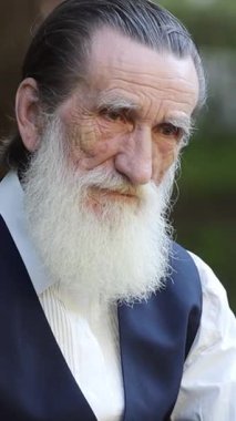 Uzun beyaz sakallı, sırtı kıvırcık saçlı, düşünceli sakin bir anda bankta oturan son sınıf öğrencisi. Dikey video.