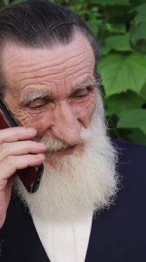 Beyaz sakallı yaşlı bir adam sık yeşil yaprakların yanında akıllı telefonuyla konuşuyor ve dışarıda demir parmaklıklar var. Dikey video.