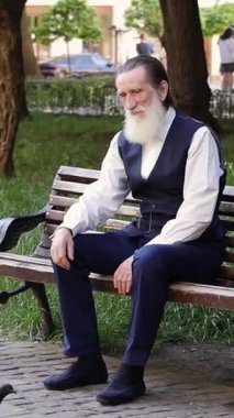 Uzun beyaz sakallı yaşlı bir adam parktaki yürüyüş yolunun yanındaki bankta tek başına dinleniyor. Dikey video.