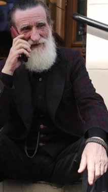 Beyaz sakallı arkadaş canlısı son sınıf öğrencisi dışarıda merdivenlerde otururken gülümsüyor ve telefonda konuşuyor. Dikey video.