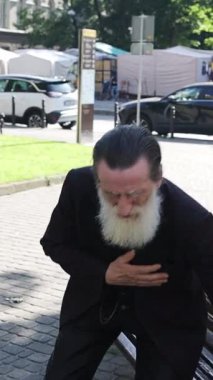 Beyaz sakallı, gülümseyen, telefonda konuşan ve parktaki bankta otururken doğal bir şekilde el kol hareketi yapan son sınıf öğrencisi. Dikey video.