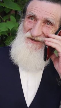 Uzun beyaz sakallı yaşlı adam şehir kaldırımında yemyeşil çalıların yanında dikilirken cep telefonuyla konuşuyor. Dikey video.