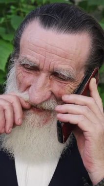 Uzun beyaz sakallı yaşlı bir adam sık yeşilliğin yanında cep telefonuyla konuşuyor ve bir ara sokakta pencere var. Dikey video.