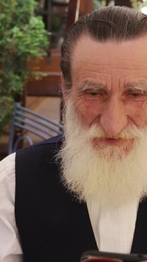 Şehirdeki bir kafede otururken akıllı telefonunu kullanan uzun beyaz sakallı yaşlı bir adam. Dikey video.
