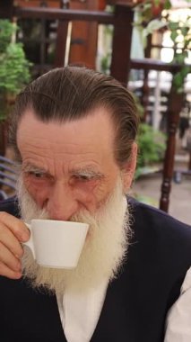 Açık bir kafede akıllı telefonunu kullanarak kahve içen uzun beyaz sakallı yaşlı bir adam. Dikey video.