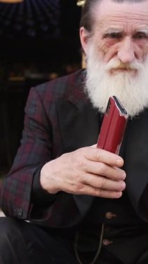 Uzun beyaz sakallı yaşlı bir adam taş basamaklarda oturuyor ve sakin bir anda elinde kırmızı bir şapka tutuyor. Dikey video.