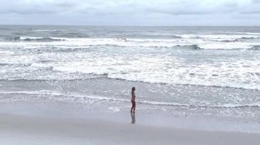 Uzun saçlı genç bir kadın bulutlu bir günde sahilde yürüyüş yapıyor.