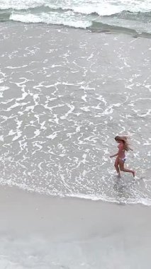 Dalgalar sahili yıkarken kumsalda yalınayak koşan genç bir kadının hava görüntüsü.