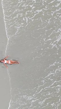 Beyaz bikinili güzel bir kadın kumlu bir sahilde uzanıyor ve okyanus dalgaları onu nazikçe yıkıyor.