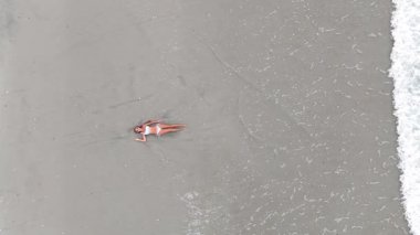 Okyanus dalgaları yavaşça kıyıya yaklaşırken bikinili genç kadın kumda dinleniyor.