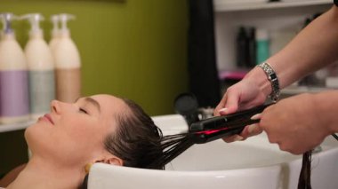 Genç bir kadın kuaförde dinlenirken kuaför keratin tedavisini ütüyle uygular.