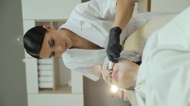 Kadın güzellik uzmanı, bir müşterinin yüzünde gençleştirici cilt tedavisi için özel bir cihaz kullanıyor.